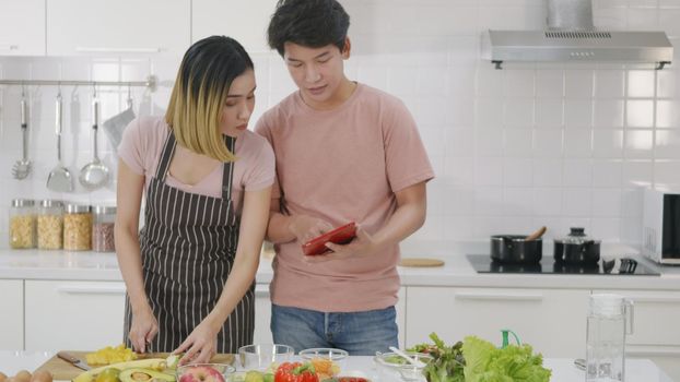 Happy Asian beautiful young family couple husband and wife cooking vegetable salad in kitchen together at home. The man and woman according online cooking class to recipe on laptop computer