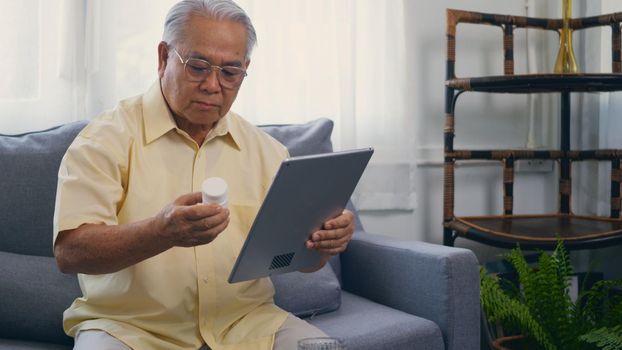 Asian elderly patient video call by digital tablet to doctor for inquire about which bottle of medication pills should be taken in living room at home,  Senior old man technology online healthcare