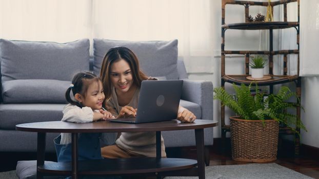 Asian young mother with laptop computer teaching her kid to learn or study online in living room at home, Mum and little preschool daughter learning online on computer, family homeschooling online