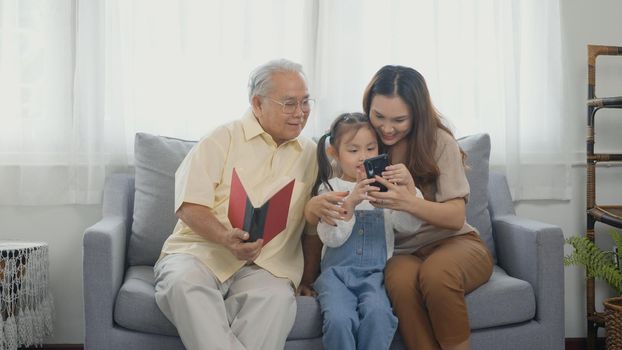 Happy family time relax. Asian grandparents laughing taking selfie with granddaughter on sofa at home by mobile smartphone. senior man or grandpa, daughter and girl kid in living room together