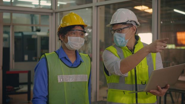 Two factory industrial workers technician or engineer worker and manager wear uniforms, safety helmets talking about production plans new project while walking at workshop manufacturing