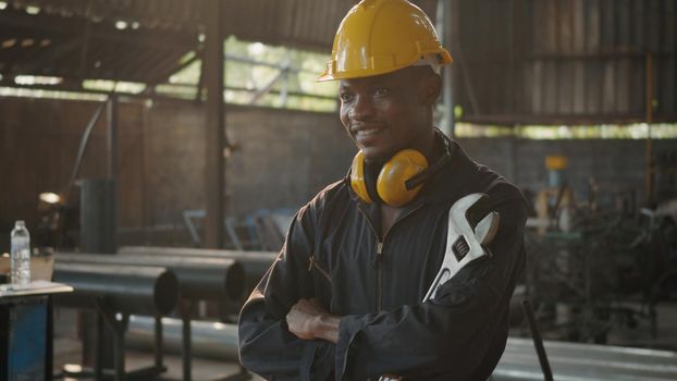 Portrait American industrial black young worker man smiling with yellow helmet in front machine, Engineer standing holding wrench tools and arms crossed at work in the industry factory.