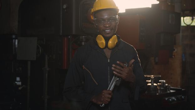 Portrait American industrial black young worker man smiling with helmet and ear protection in front machine, Engineer standing holding wrench on his hand at work in industry factory.