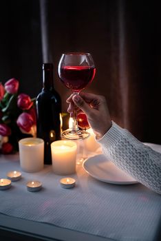 Romantic Candlelight Dinner for Two Lovers. Set of tableware with bouquet of tulips, wine and candles, dark background. Woman hand holding a glass of wine