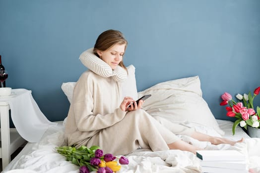 Happy woman sitting on the bed wearing pajamas typing a message on the phone, blue wall background