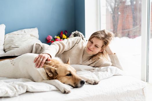 Happy young woman lying in the bed with her dogs, blue wall background