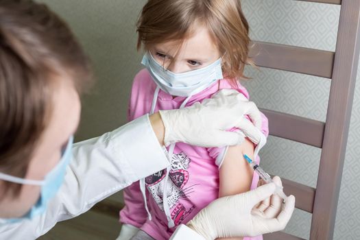 Little caucasian girl in a medical mask sits on a chair in the doctor's cabin and receives a vaccination, looks at the doctor