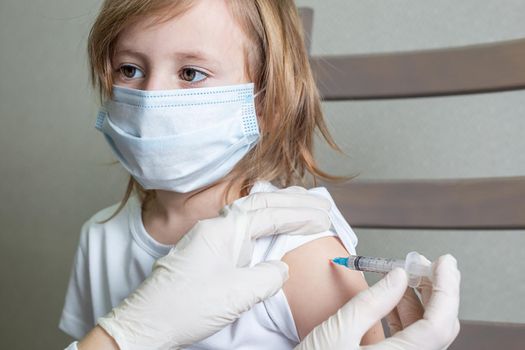 Little caucasian girl in a medical mask sits on a chair in the doctor's cabin and receives a vaccination, looks at the doctor