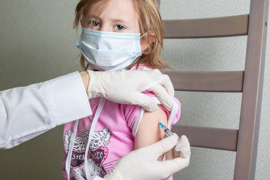 A 5-year-old Caucasian girl in a medical mask receives a vaccine against a coronavirus infection, looks at the camera.