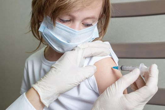 A 5-year-old Caucasian girl in a medical mask receives a vaccine, peeping with eyes down on the syringe