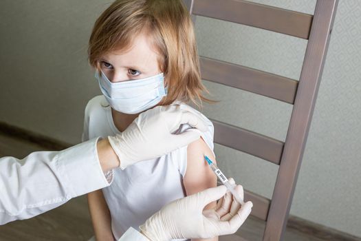 Doctor in a medical mask and gloves makes an injection of a vaccine against a coronavirus infection into the shoulder of a little caucasian girl in a medical mask, the girl looks at the doctor