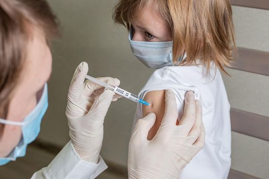 Doctor in a medical mask and gloves makes an injection in the shoulder of a little caucasian girl in a medical mask, the girl looks at the doctor