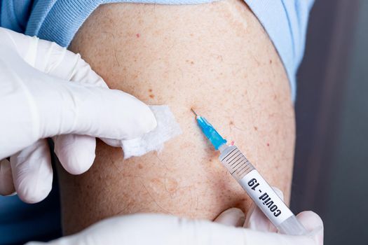 An elderly man of Caucasian appearance in a mask receives a dose of coronavirus vaccination at home, the doctor came to the patient to give a protective injection against the disease