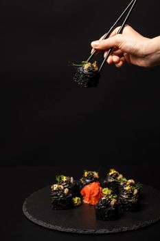 Hand with chopsticks holds a sushi. Custom sushi roll with black rice, crab meat, avocado, smoked salmon mousse, oar caviar, masago, shrimp cocktail, edible gold leaf, ginger, wasabi on black table