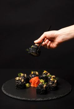 Hand with chopsticks holds a sushi. Custom sushi roll with black rice, crab meat, avocado, smoked salmon mousse, oar caviar, masago, shrimp cocktail, edible gold leaf, ginger, wasabi on black table