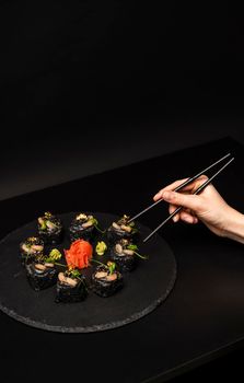 Hand with chopsticks wants to take custom sushi roll with black rice, crab meat, avocado, smoked salmon mousse, oar caviar, masago, shrimp cocktail, edible gold leaf, ginger, wasabi on black table