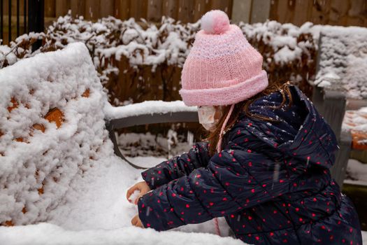 Portrait of a cute baby girl in pink hat playing in the snow wearing a face mask