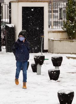 A cute boy wearing a black hat and a blue face mask walking in the snow