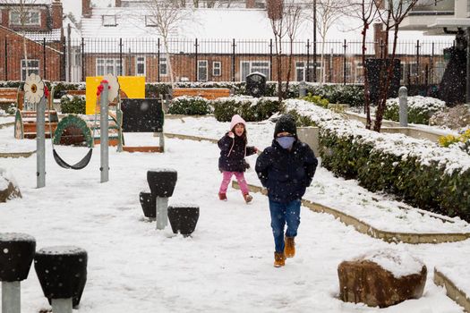A cute baby girl wearing a blue jacket and a pink hat and a cute boy wearing a black coat and hat and blue mask in a snowy playground