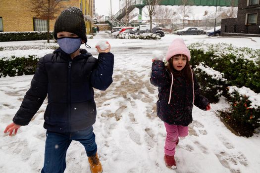 A cute boy wearing a black hat and a blue mask and a girl wearing a a pink hat thowing snow balls in the street