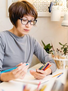 Woman with short hair cut is drawing in notebook. Calming hobby, antistress leisure. Artist at work. Cozy workplace.