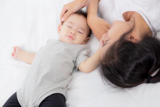 Young asian mother touch head of little baby girl with tender on bed in the bedroom, mom love newborn and care, woman with expression with child together, parent and daughter, family concept.