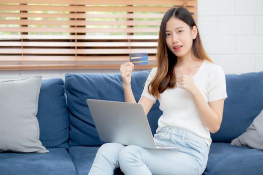 Young asian woman sit on sofa using laptop computer shopping online with credit card buying to internet, happy girl payment with e-business on couch, purchase and payment, business concept.