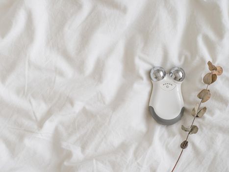 Microcurrent concept. Face massager with micro currents. Home use device for anti-aging and face lifting on white bed. Top view or flat lay, copy space for text.
