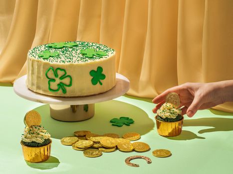 St Patrick's Day food concept. Modern still life with sweet food for Saint Patrick's Day party with female hand. Cake decorated shamrocks, green velvet cupckakes, chocolate golden coins and horseshoe