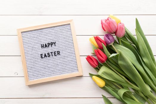 Spring and Easter concept. Bouquet of colorful tulip flowers and letter board with the words Happy Easter top view flat lay on white wooden background