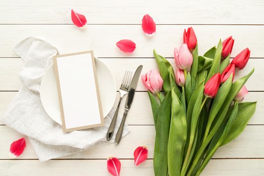 Festive wedding, birthday, romantic table setting with silver cutlery and fresh pink tulips bouquet. Blank menu card mockup. Restaurant menu concept. Flat lay, top view