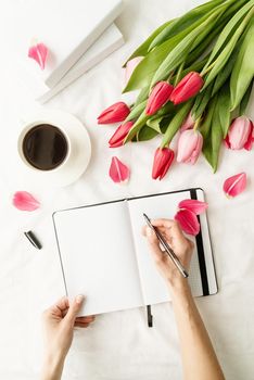 Holiday and celebration. Spring holidays. Woman hand making notes in opened notebook, decorated with tulips, coffee cup and books, top view flat lay