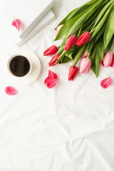 Holiday and celebration. Top view of pink tulips, coffee cup and books on white bed, flat lay