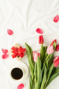 Holiday and celebration. Top view of pink tulips, coffee cup and gift box on white bed, flat lay