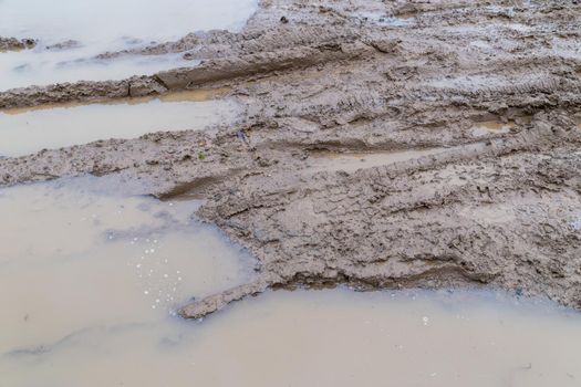 dirty wet clay mud road with tire tracks and puddles at day time - full frame background with hard sun light