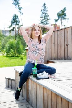 A young girl of 20 years old Caucasian appearance enjoys the sun and weather while sitting on a wooden podium in the park on a summer day.The girl is dressed in a floral T-shirt and jeans