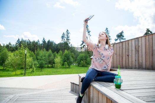 A young girl of 20 years old Caucasian appearance makes a selfie on her mobile phone while sitting on a wooden podium in the park on a summer day.The girl is dressed in a floral T-shirt and jeans, 