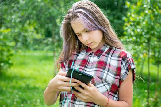 A young girl of 15 years old Caucasian appearance wary with apprehension looks into her mobile phone on the lawn in the park on a summer day. The girl is dressed in a plaid shirt and jeans.