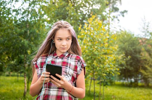 A young girl of 15 years old Caucasian appearance looks around and holds a mobile phone in her hands in the park on a summer day. The girl is dressed in a plaid shirt and jeans.