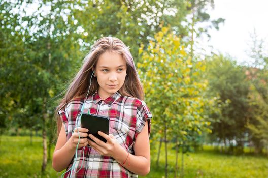 A young girl of 15 years old Caucasian appearance looks around and holds a mobile phone in her hands in the park on a summer day. The girl is dressed in a plaid shirt and jeans.