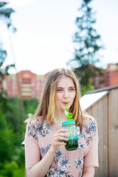 A young girl of 20 years old Caucasian appearance enjoys the sun and weather and drinks smoothies from a large glass while sitting on a wooden podium in the park on a summer day.