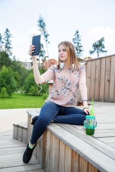 A young girl of 20 years old Caucasian appearance makes a selfie on her mobile phone while sitting on a wooden podium in the park on a summer day.The girl is dressed in a floral T-shirt and jeans,