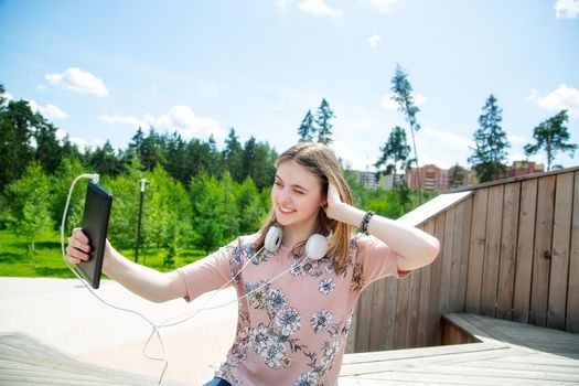 A young girl of 20 years old Caucasian appearance makes a selfie on her mobile phone while sitting on a wooden podium in the park on a summer day.The girl is dressed in a floral T-shirt and jeans, 