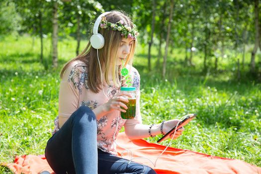 A young girl of 20 years old Caucasian appearance listens to music on her mobile phone and drinks smoothies from a large glass, sitting on the lawn in the park on a summer day. 