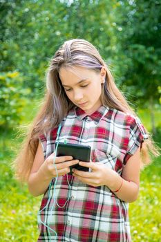 A young girl 15 years old Caucasian appearance reads and writes a message in a mobile phone in a park on a summer day. The girl is dressed in a plaid shirt and jeans.