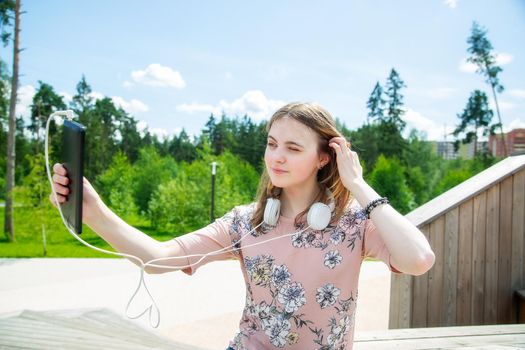 A young girl of 20 years old Caucasian appearance makes a selfie on her mobile phone while sitting on a wooden podium in the park on a summer day.The girl is dressed in a floral T-shirt and jeans, 