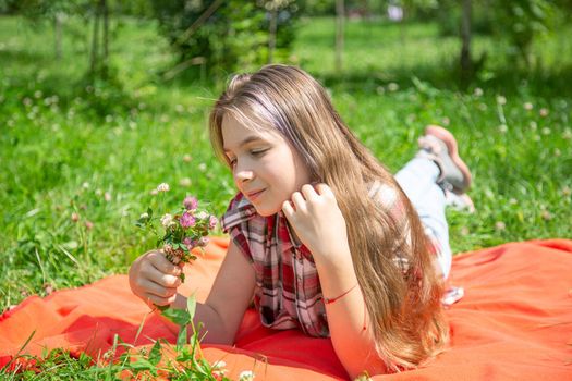 A young girl of 15 years old Caucasian appearance enjoys the sun and the weather and a bouquet of wild flowers lying on the lawn in the park on a summer day. The girl is dressed in a plaid shirt 
