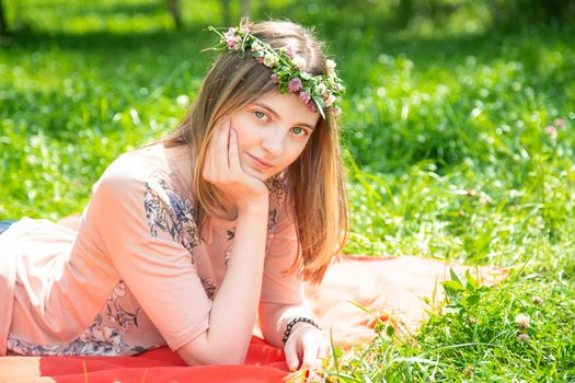 Young girl 20 years old Caucasian appearance smiling looking at the camera while lying on the lawn in the park on a summer day. The girl is dressed in a T-shirt and jeans and a wreath of wildflowers.