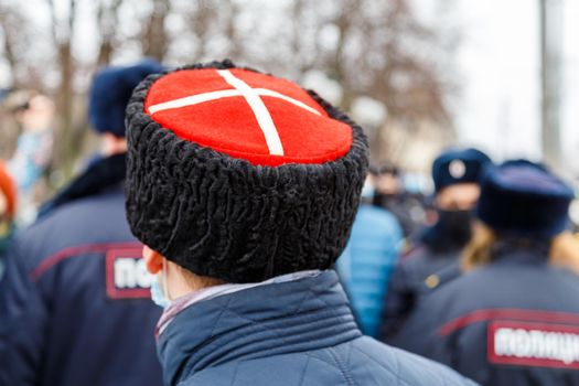 man in cossack hat with white cross on red watching blurry crowd of people - view from back.