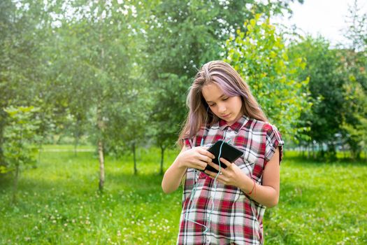 A young girl of 15 years old Caucasian appearance wary with apprehension looks into her mobile phone on the lawn in the park on a summer day. The girl is dressed in a plaid shirt and jeans.